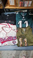 Four sportswear items laid out: Phillies white baseball jersey with red piping, black Phillies t-shirt, green Eagles jersey with number 11, green Eagles camouflage number 11 jersey.