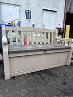 Front view of beige plastic outdoor storage bench showing wood grain texture, vertical slat backrest, hinged dark gray seat and armrests. Dust on surface visible.