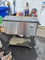 Front view of a metal portable rolling cooler with dual black lids, bottle opener on the front, supported on caster wheels with a lower shelf for storage. Cooler presents some dust and outdoor wear.