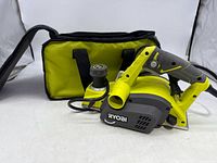 RYOBI electric planer laying in front of a yellow and black carrying bag on white background