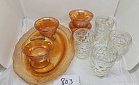 Photo showing four Marigold Carnival glass sherbet cups with pedestal bases arranged on matching Marigold oval tray/platter. Also includes four clear iridescent thumbprint textured glasses with irregular bubble indentations.