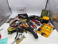 Overview photo showing various hand tools scattered including hammers, pliers, a tape measure, screwdrivers, and bit sets
