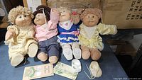 Four vintage Cabbage Patch Kids dolls seated together, showing overall condition, clothing, and included accessories.