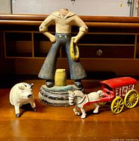 Side view of all three vintage cast iron toy items on wooden surface under cabinet: Popeye door stopper missing head, small white pig bank, and red horse-drawn ice wagon.