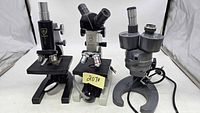 Front view of three vintage microscopes including Spencer, Bausch & Lomb, and Parco models with varying eyepiece setups and body colors.