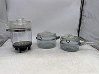 Full lot photo showing 6-cup glass coffee percolator on left with black stand and two glass saucepan pots with lids on right