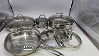 Set of Wolfgang Puck stainless steel cookware pieces arranged showing various pots, pans, and lids