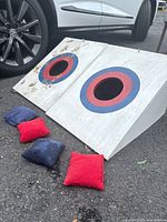 Two wooden bean bag toss boards with circular red and blue target areas and four bean bags, placed on ground beside a vehicle.
