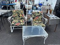 Two white wrought iron chairs with floral cushions, and a matching wrought iron and glass coffee table shown from the front, outdoor setting.