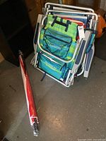 Stack of three folded backpack beach chairs showing aluminum frames, striped fabrics, storage pouches, shoulder straps; umbrella beside