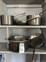 Full view of the cookware set displayed on shelf showing three saucepans, one stock pot, and one frying pan with metal handles and stainless steel interiors.
