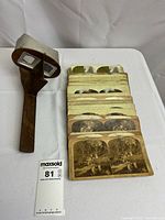 Stereoscope viewer with wooden handle and white metal/enamel hood placed on table next to stacked vintage stereoview photo cards.