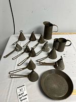Image shows various antique metal kitchen utensils including 11 metal funnels with handles and loops, two metal pitchers, and a shallow metal plate, arranged on a white cloth.