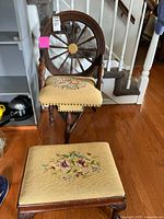 Front angle view of antique wooden chair with wheel back design and floral tapestry seat. Matching footstool in front with floral upholstery and visible broken leg.