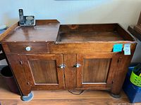 Full front view of the antique wooden dry sink showing top surface, drawer, and two doors below with metal handles and white knob on drawer.