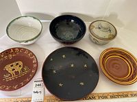 Photo showing six ceramic pottery items arranged on white surface with ruler for scale. Items include three bowls, three plates with various hand-painted and glazed designs.