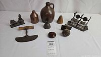 Overview of all items arranged on white surface including stoneware jug, small bottles, metal blade tool, wooden paperweight, brass door knob, metal ink blotter, ceramic funnel, and double inkwell set