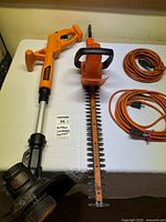 Photo showing Black+Decker orange weed wacker, hedge trimmer, and orange extension cord on table.
