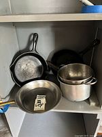 Shelf with two cast iron frying pans, stainless steel frying pan with label, stainless steel colander, and stainless steel saucepan, all showing visible use and residue.