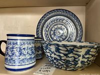 Photo shows two blue and white ceramic mugs with foliage pattern, and a matching large platter and bowl behind them.