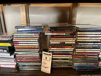 Photo of stacked CDs and cassette tapes, showing numerous jewel cases and cassette cases in mixed condition, showcasing the volume and variety of the collection.