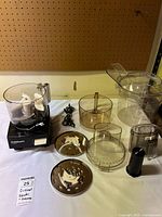 Photo showing all Cuisinart food processor parts and accessories arranged on a table with pegboard background. Items include base motor unit, various clear plastic bowls and lids, blades, discs, and cords.