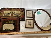 Two decorative hooked rugs laid flat, showing sheep and floral designs with ruler for scale. Two small framed floral needlepoint artworks and oval wood framed mirror next to rugs.