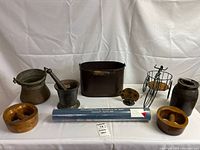 Overall lot showing all included items arranged on a white backdrop: brass and cast iron mortar and pestle, two wooden herb choppers, metal bin and bucket, ceramic jug, wire figure tricycle, metal hedgehog sculpture, and blue-rolled exhibition poster.
