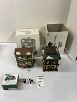 View of two Department 56 Dickens' Village ceramic buildings and two Victorian-style figurines with their original boxes placed behind them on a white table.