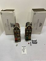 Two Dept. 56 Christmas In The City Heritage Village buildings with original boxes placed behind them; various small accessories placed in front.