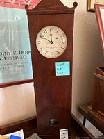Front view of tall wooden wall clock showing the clock face, brand inscription, and decorative top molding.
