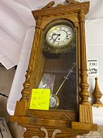 Front view of wooden Emperor wall clock showing face, gold detailing, pendulum, and detached finials on right side.