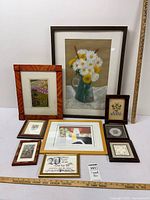 Photo showing the entire lot of 10 framed art prints arranged and leaning against a white surface; prints include floral, scenic, and folk art styles in various wood and metal frames.