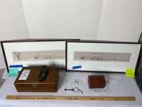 Photo showing two framed art prints with Amish winter scenes, two wooden boxes, metal shoe figurine, magnifying loupe, and eyeglasses laid out with a ruler for scale.