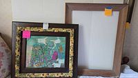 Photo showing two framed pieces leaning against a wall, one visible with an ornate floral-painted wood frame containing a folk art painting with angels and flowers.