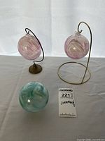 Three hand-blown glass ornaments: two pink swirled glass balls displayed on metal stands (one darker metal, one brass-colored), and one green/blue swirled glass ball sitting on surface.