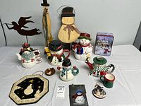 Full view of all holiday decor items arranged on a white surface showing snowman themed ceramics, wood, and tin pieces including ornaments and a metal angel silhouette