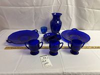 Eight cobalt blue glassware pieces arranged on white backdrop including vase, handled plates, divided dish, small cup, creamer, and sugar bowl.