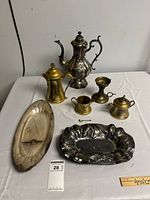 Photo showing full lot including silverplate teapot, brass coffee pot, sugar bowl, creamer, small spoon, open dish/compote, oval tray, and rectangular corn motif platter on white surface.