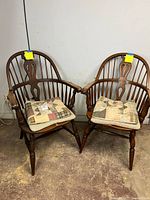 Pair of medium brown traditional Windsor wood arm chairs each with quilted patchwork seat cushions, shown in a basement setting.