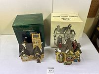 Two porcelain lighted houses with their original boxes. One box is green and the other white and damaged. The lighting cords, bulbs, or plugs are not visible.