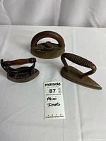 Set of three antique miniature irons placed on white cloth with a label showing lot number and description.