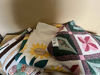 Three handmade quilts stacked showing the sunflower quilt center, geometric pink-green quilt, and a striped patchwork quilt beneath