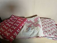 Photo showing three folded quilts in red, white, and pink colors with various patterns placed flat against a cream wall.