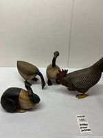 Four wooden animal figurines displayed on white surface: black and tan rabbit, black and tan geese one with head up and one feeding, and detailed painted chicken with red comb.