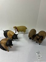 Five wood and resin rustic animal figurines including two pigs, one cream sheep with scarf, and two brown rams arranged on white surface.