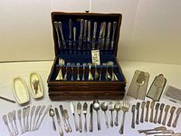 Full view of wooden flatware case with blue velvet lining holding various knives, forks, spoons laid out inside and more pieces displayed around case.
