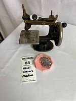 Miniature Singer sewing machine with visible rust and wear, metal parts including the needle area. Small plastic container with colorful pins beside it. White background with a label showing lot number and description.