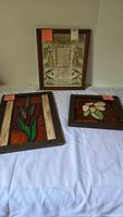 Three framed decorative panels viewed on a white cloth: one framed print artwork with angels and birds in center; two stained glass art panels with orange backgrounds and floral designs on left and right.