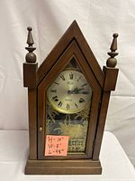 Front view of wooden mantel clock with triangular top, glass door with Roman numeral dial, date window and decorative gold detailing, wooden finials on top corners, showing surface wear noted on base and top.
